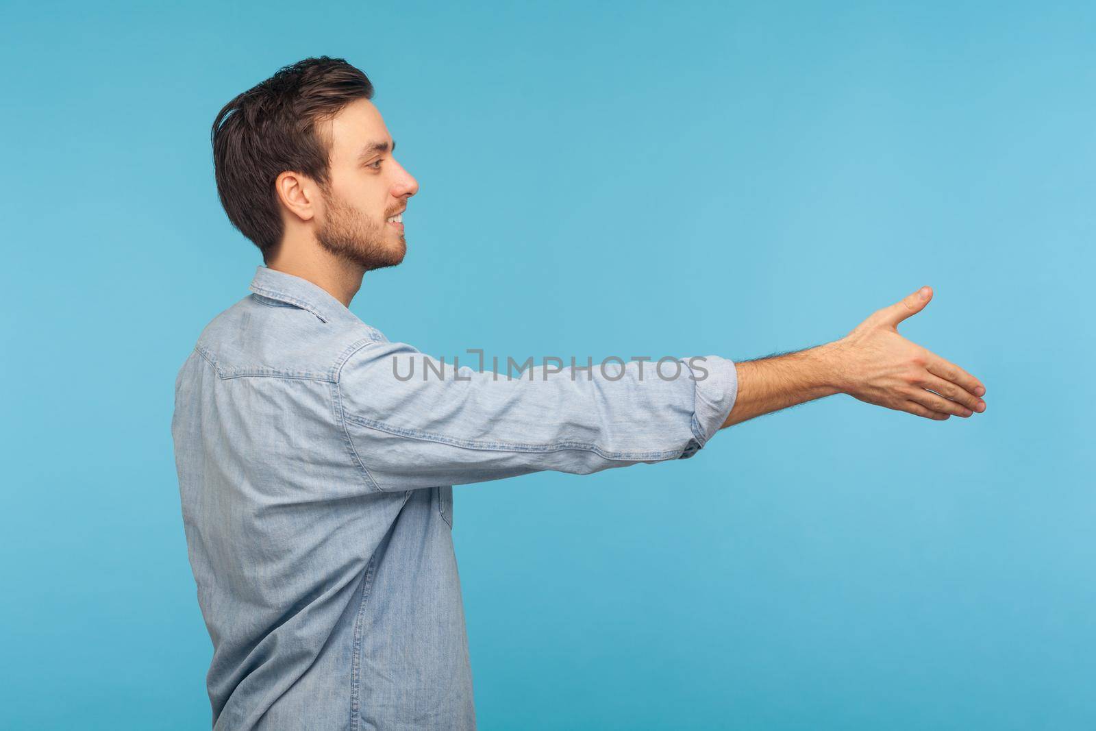 Hi, nice to meet you. Side view of friendly smiling handsome man applicant in worker denim shirt outstretching hand, offering handshake on job interview. indoor studio shot isolated on blue background