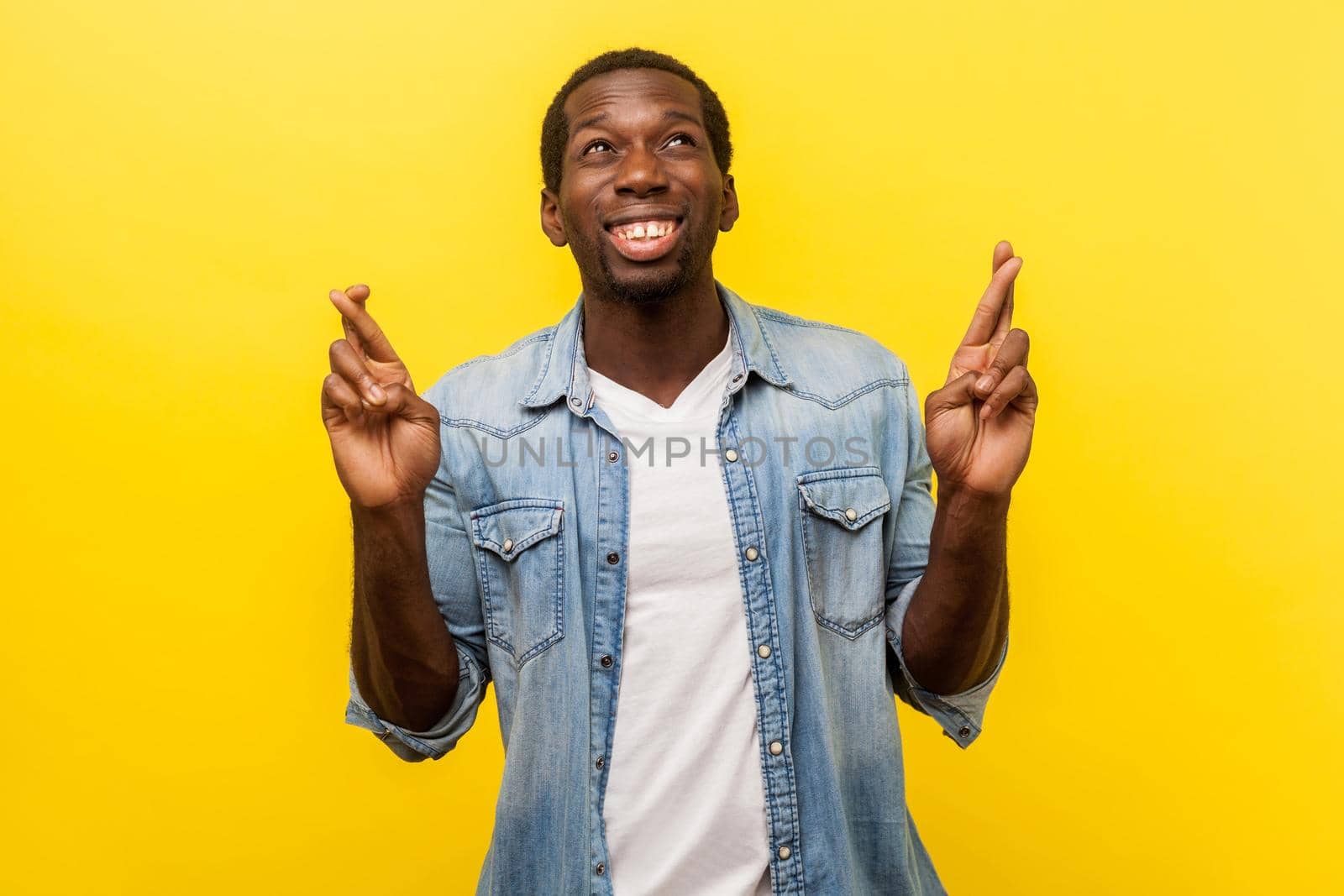 Hope for good luck. Portrait of young man in denim casual shirt with rolled up sleeve crossing his fingers and looking up with eyes full of hope, wishing fortune. indoor studio shot isolated on yellow