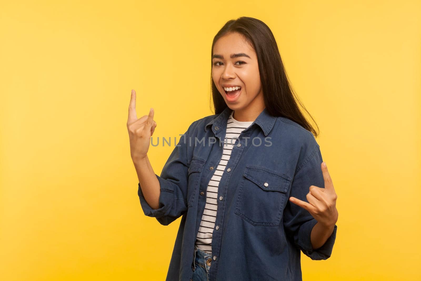 Yeah, that's wonderful. Portrait of joyful girl in denim shirt feeling crazy, showing devil horns, rock and roll hand gesture, delighted of success. indoor studio shot isolated on yellow background