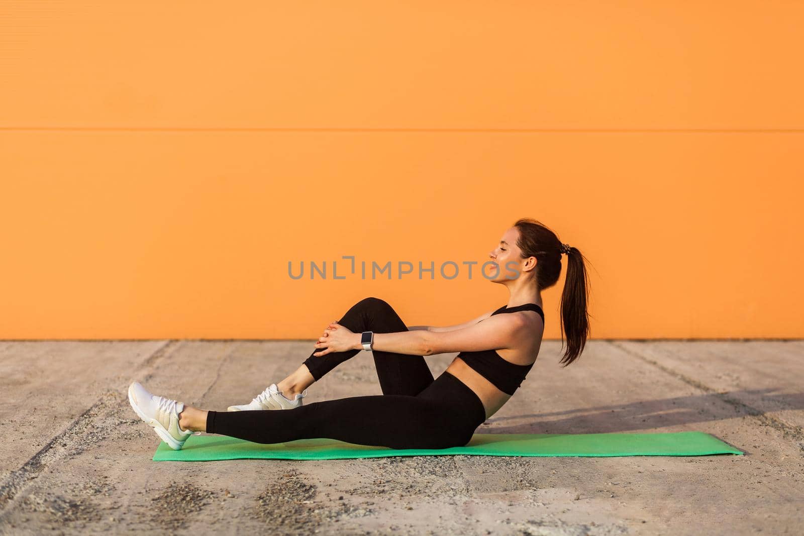 Happy smiling athletic girl in tight sportswear, black pants and top, doing aerobics gymnastics, stretching legs on yoga mat, training abs muscular and flexibility. Health care, sport activity outdoor