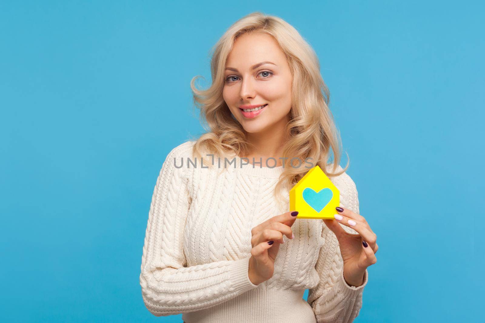 Happy smiling woman with curly blond hair holding paper house with painted heart and looking at camera, charity, shelter. Indoor studio shot isolated on blue background