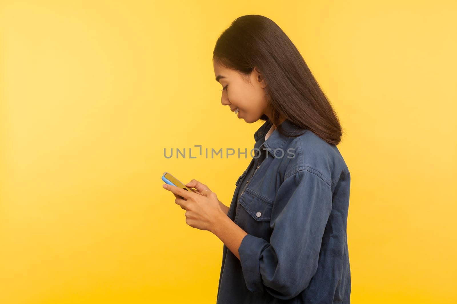 Side view of happy girl in stylish denim shirt typing message and smiling, dialing number calling on cell phone, using online mobile application, searching web. indoor studio shot, yellow background