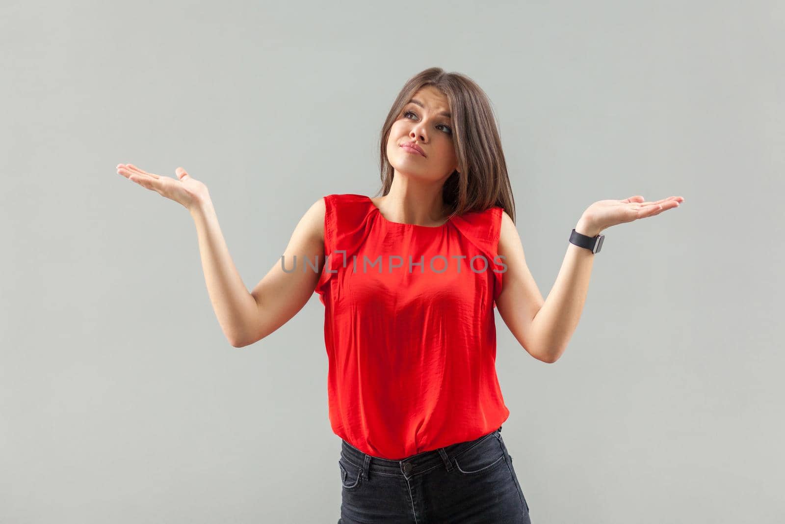 Portrait of puzzled beautiful brunette young woman in red shirt standing with raised arms, looking away thinking and confused. indoor, studio shot, isolated on gray background.