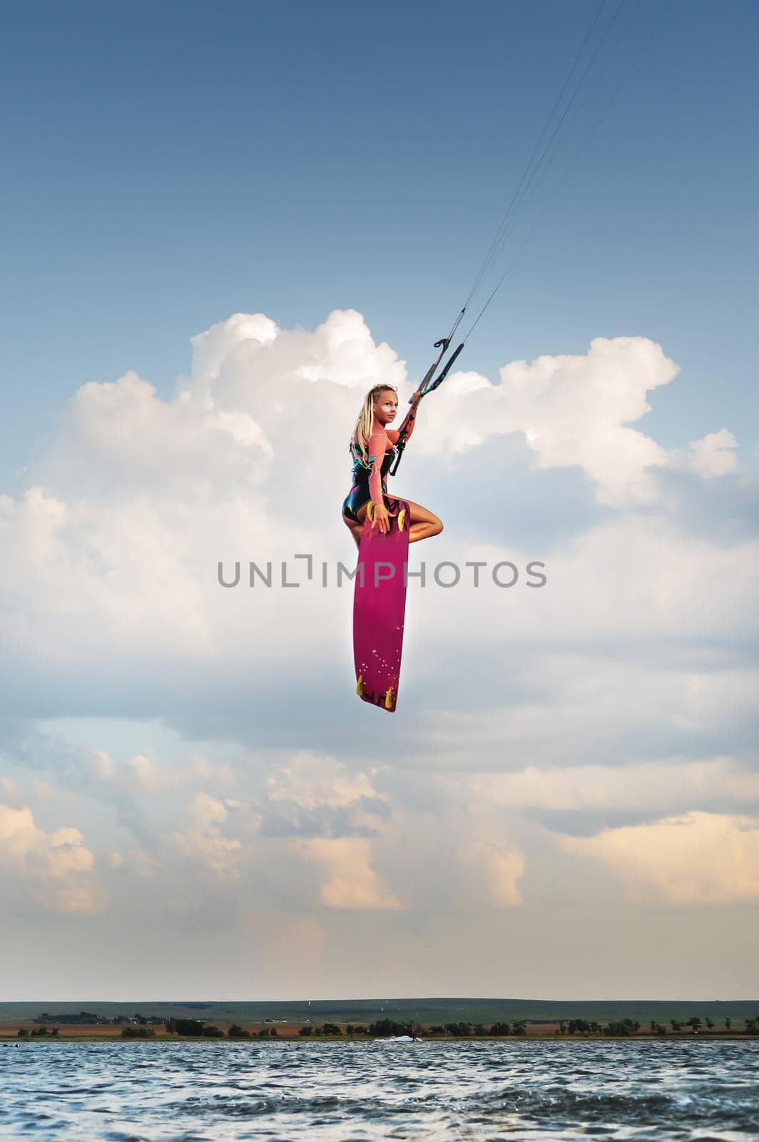 Attractive slender caucasian young woman doing kiteboarding stunt against sunset clouds and beautiful sky. Athlete makes a kitesurfing jump while looking at the camera.