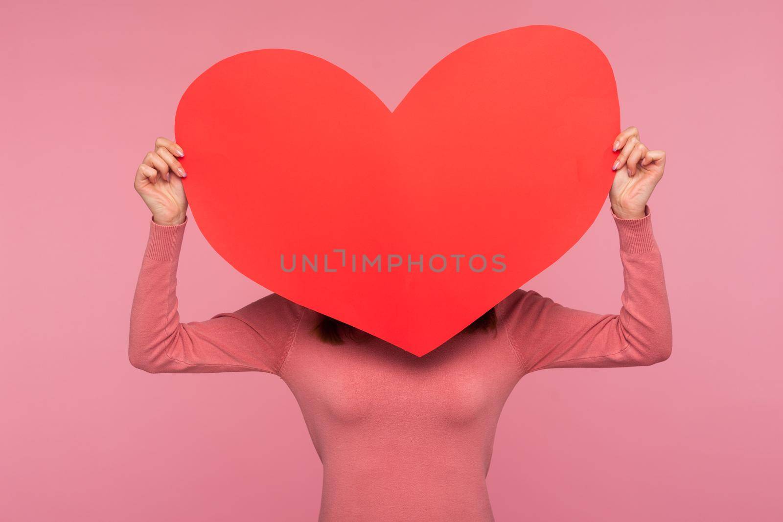 Woman in pink sweater hiding behind big red paper heart, charity concept, help and support, healthcare and transplantation organisation. Indoor studio shot isolated on pink background