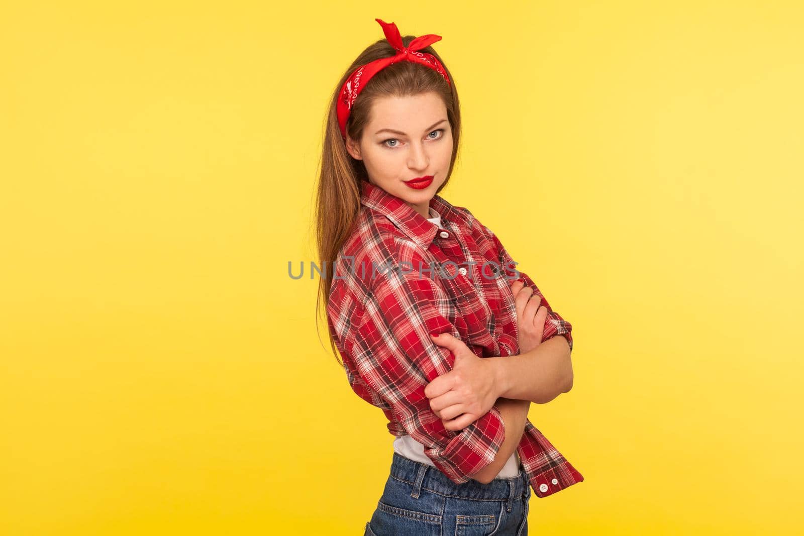 Portrait of sexy pinup girl with bright makeup, red lipstick, wearing checkered shirt and headband, standing with crossed hands and looking seductively, retro vintage 50's style. studio shot isolated