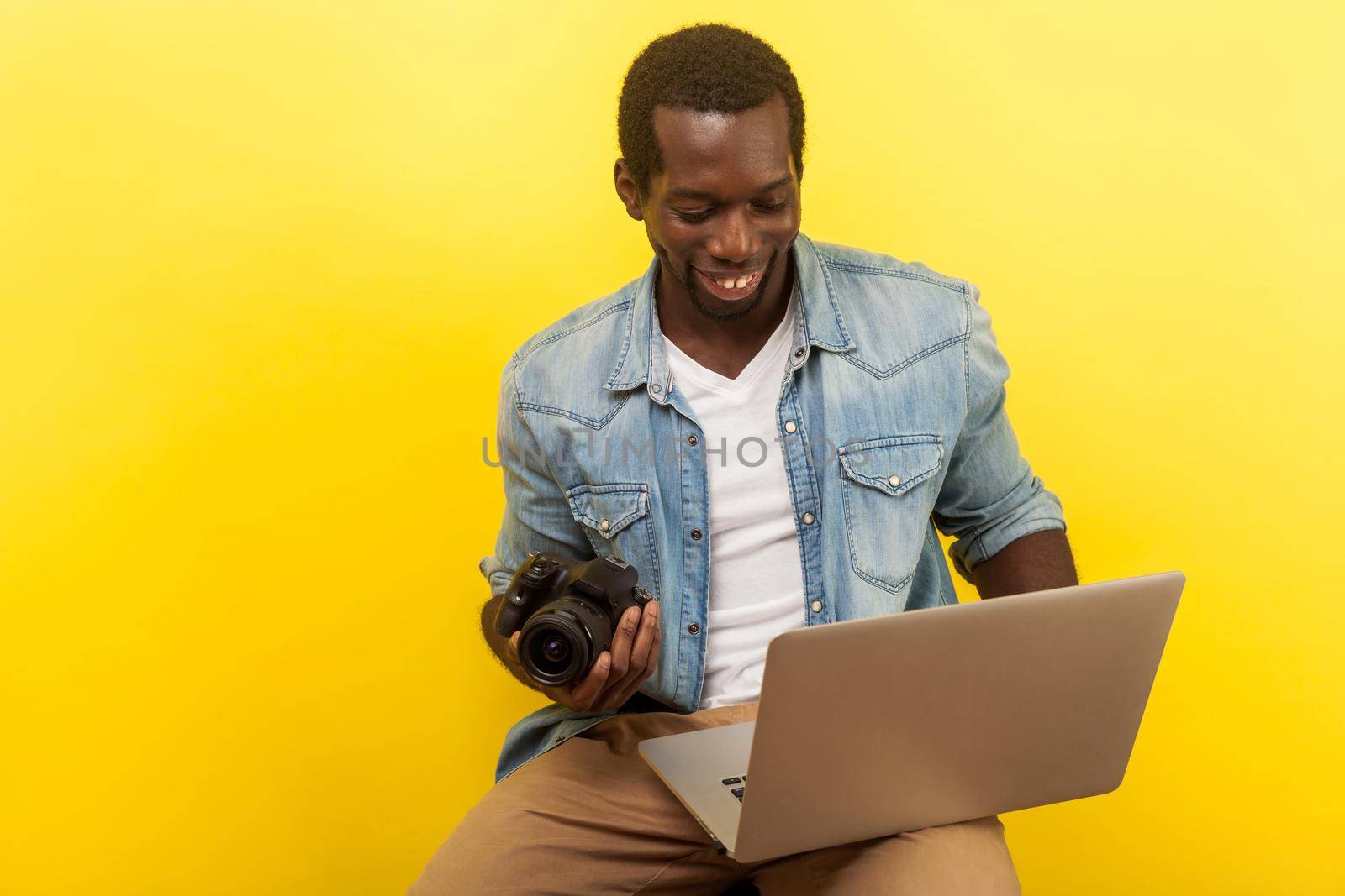 Portrait of positive male photographer in denim shirt holding digital dslr camera and laptop, going to take picture, using photo editing app on computer. studio shot isolated on yellow background