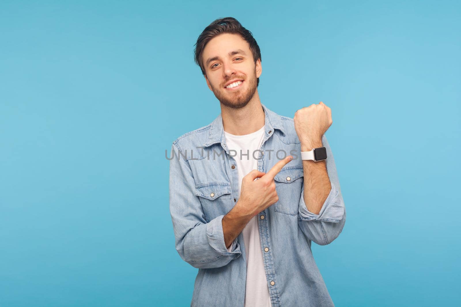 Please, hurry up. Portrait of cheerful punctual man in worker denim shirt pointing at wrist watch and smiling, showing smartwatch devise with mock up display. indoor isolated on blue background