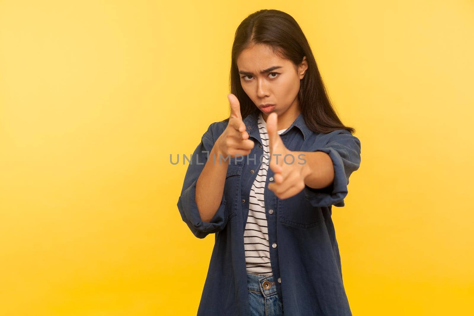 I'll shoot you. Portrait of purposeful dangerous girl in denim shirt shooting to camera with finger pistols, threatening to kill, gesturing weapon. indoor studio shot isolated on yellow background