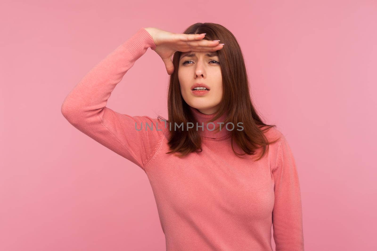 Curious brunette woman in pink sweater looking far away with hand over head, trying to see something, bad vision. Indoor studio shot isolated on pink background