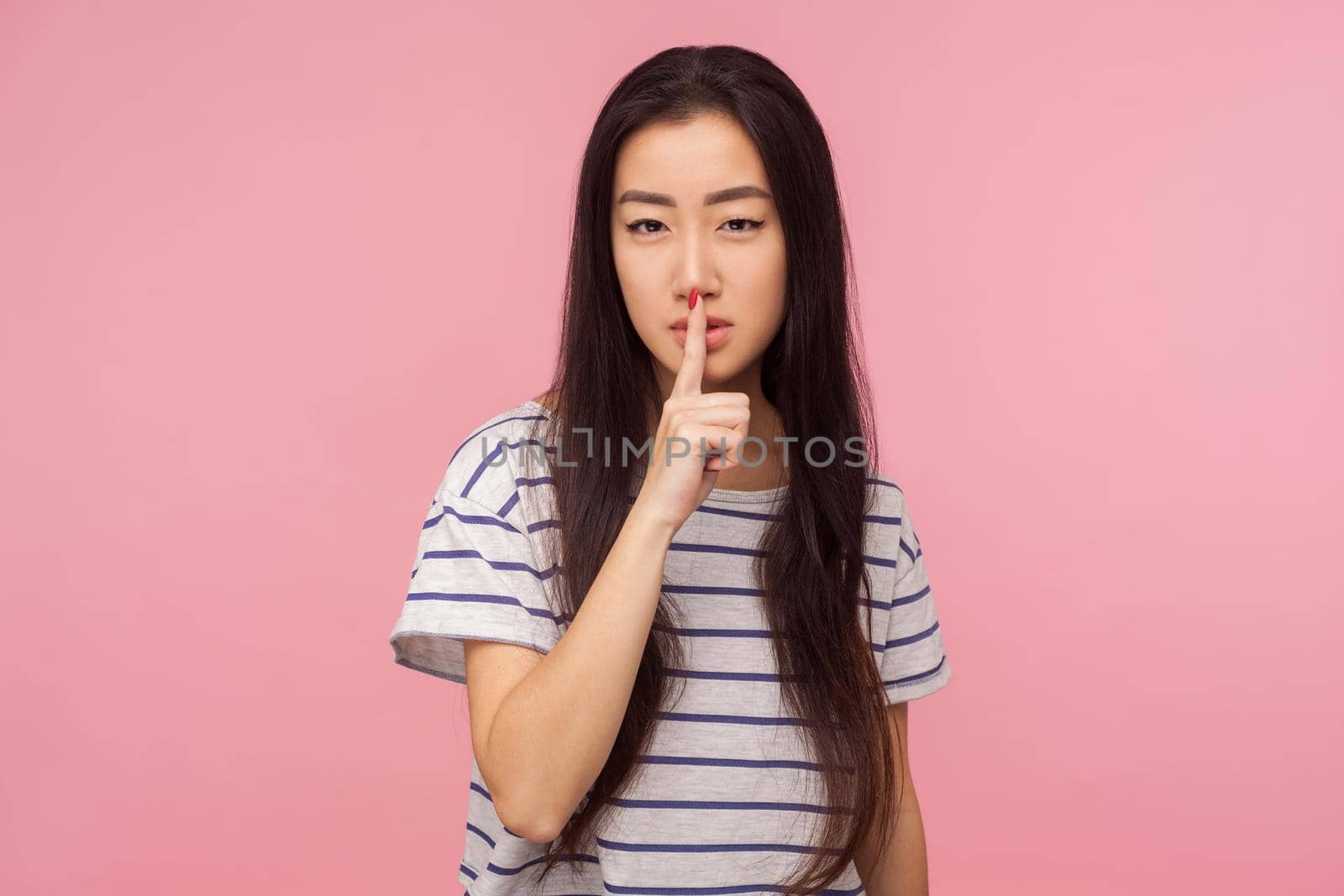 Portrait of attractive girl with long brunette hair and alluring eyes holding finger on lips, making silence gesture and asking to keep affair in secret. indoor studio shot isolated on pink background