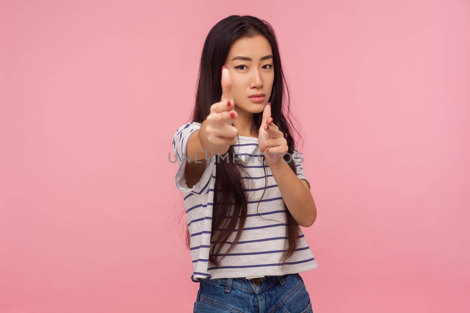 I'll kill you. Serious danger girl with brunette hair in striped t-shirt pointing finger guns to camera and shooting, gesturing weapon, aiming right at target. studio shot isolated on pink background