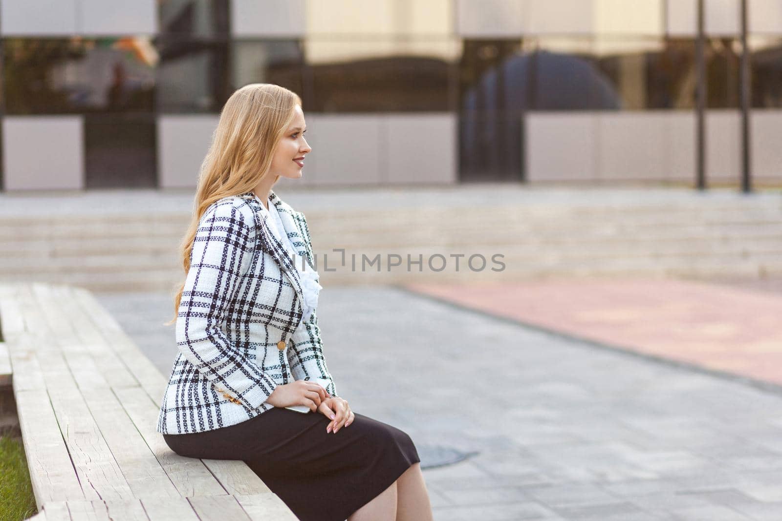 Side view of beautiful elegant successful businesswoman with long blond hair in plaid jacket and skirt sitting on bench relaxing, looking aside with cute smile. outdoors, near business center building