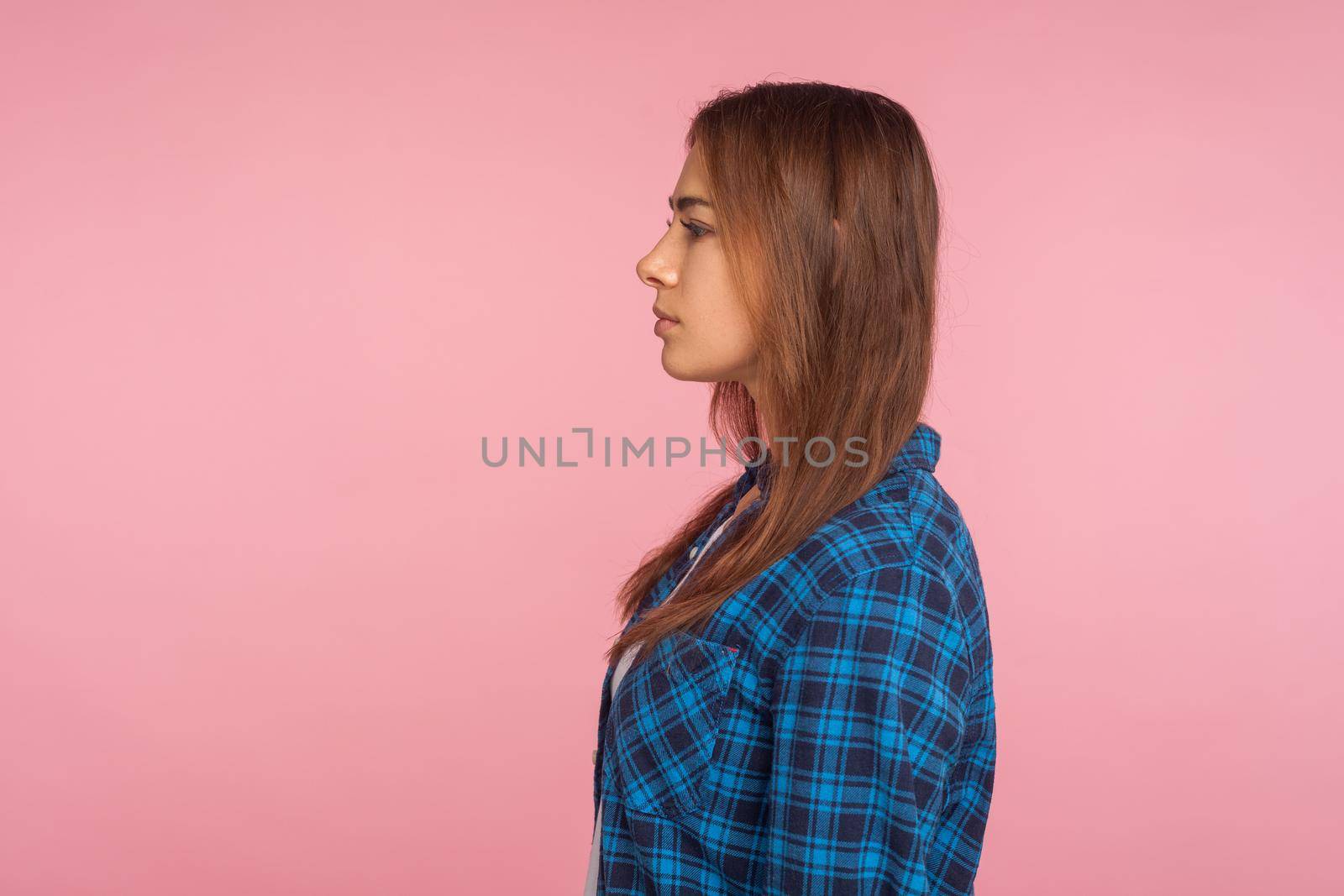 Profile of serious unsmiling girl in checkered shirt looking to side copy space with calm attentive focused expression, student or employee in casual outfit. studio shot isolated on pink background