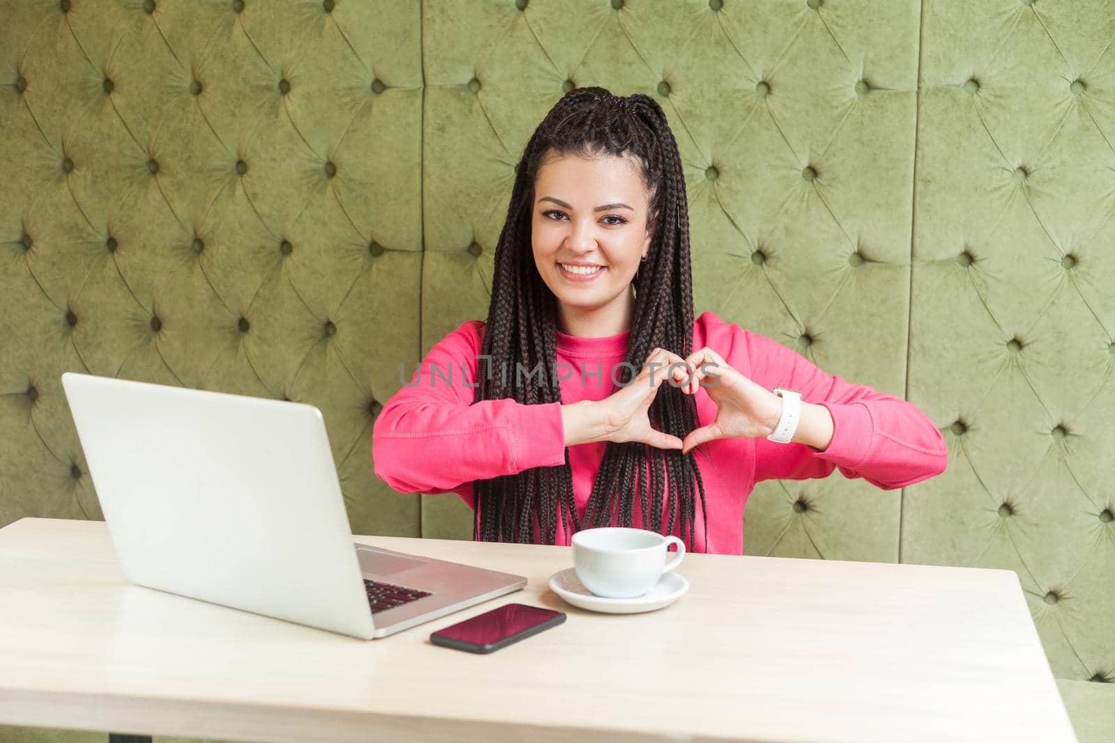 Love you. Portrait of romantic happy young girlfriend with black dreadlocks hairstyle in pink blouse sitting in cafe and showing heart shape with gesture, looking at camera with toothy smile. Indoor