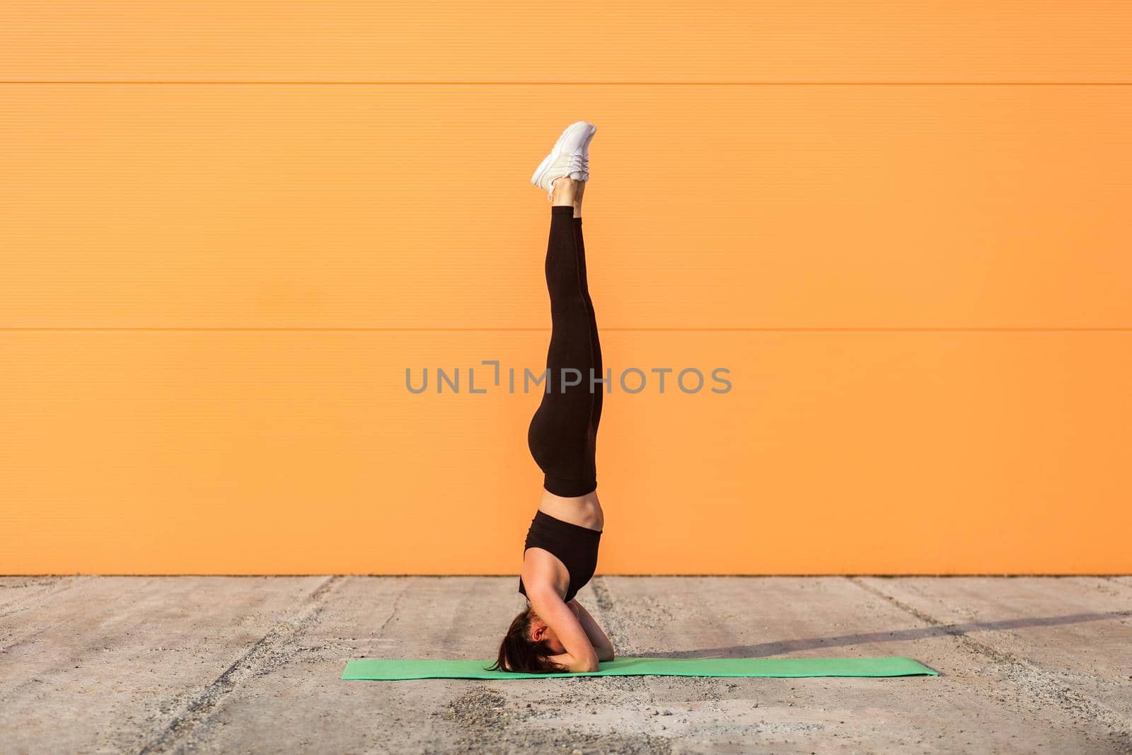 Full length girl with perfect athletic body in tight sportswear, black pants and top, doing yoga headstand pose on mat, gymnastics for body balance and flexibility. Health care, sport activity outdoor