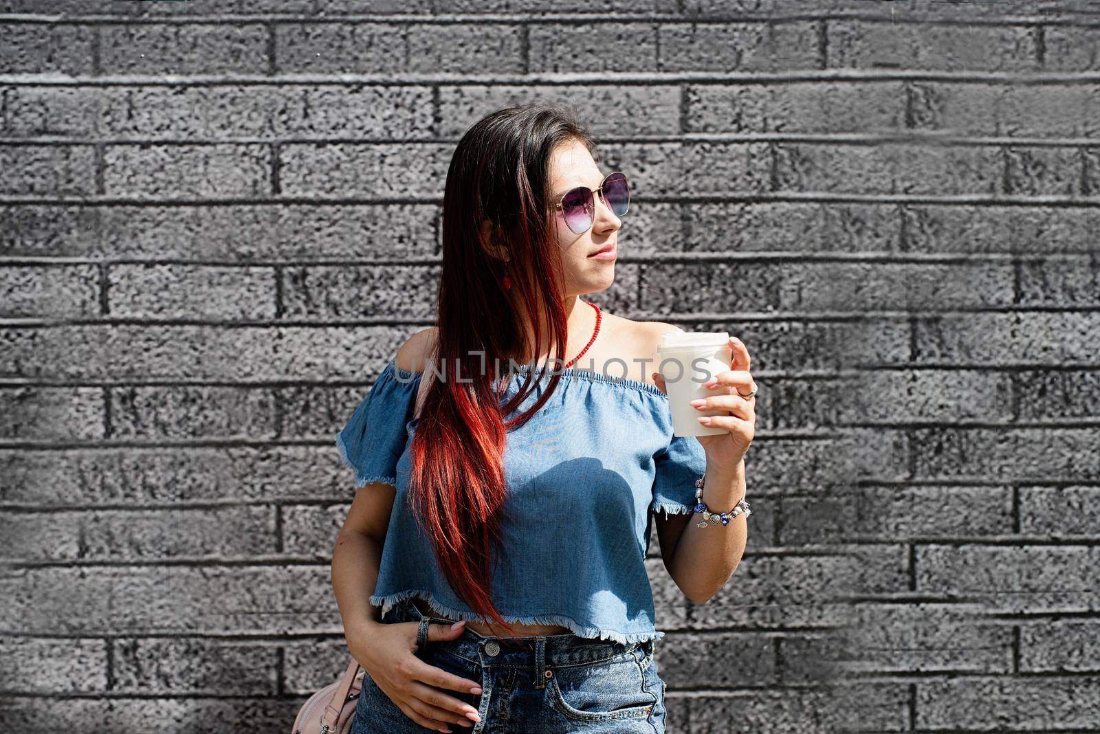 stylish young woman wearing jeans shirt, sunglasses and bag, drinking coffee to go at street, black brick wall background by Desperada