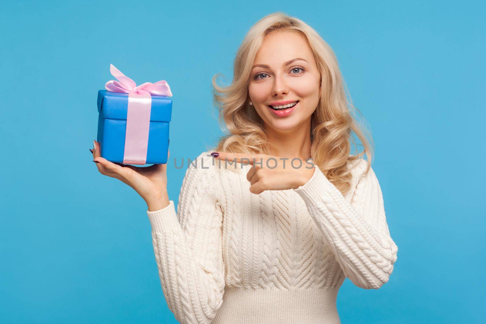 Happy satisfied blond woman pointing finger on gift box with bow she holding with toothy smile on face, present, bonuses. Indoor studio shot isolated on blue background