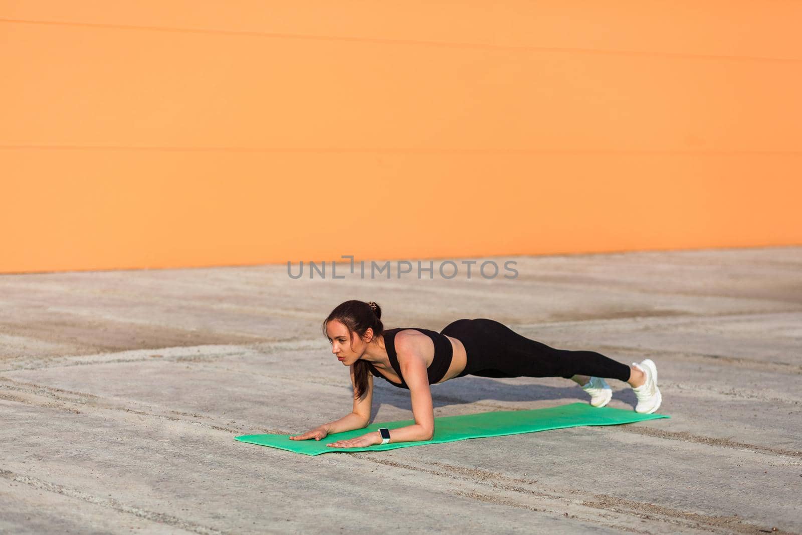 Fit slim brunette woman in tight sportswear, black pants and top, practicing yoga, standing in Ardha Phalakasana low plank pose, training muscles and strength. Health care, sport activity outdoor