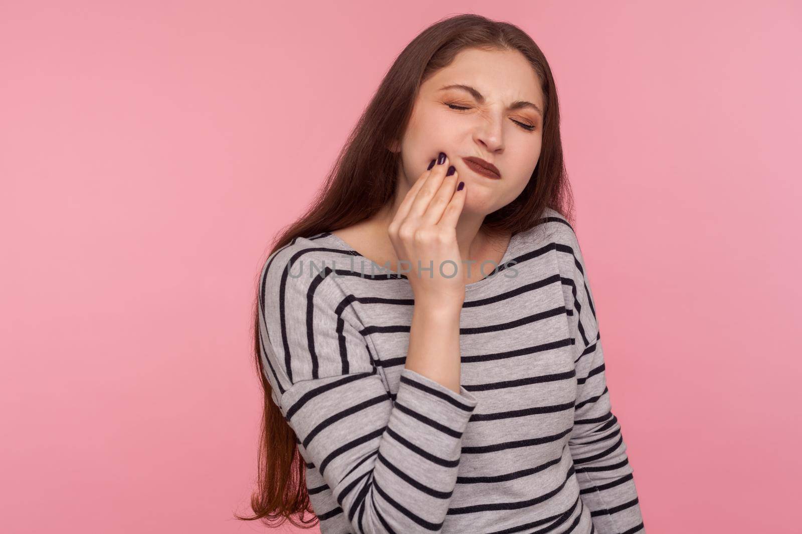 Dental problems. Portrait of unhappy sick woman in striped sweatshirt touching sore cheek, frowning suffering toothache, cavities or sensitive enamel. indoor studio shot isolated on pink background