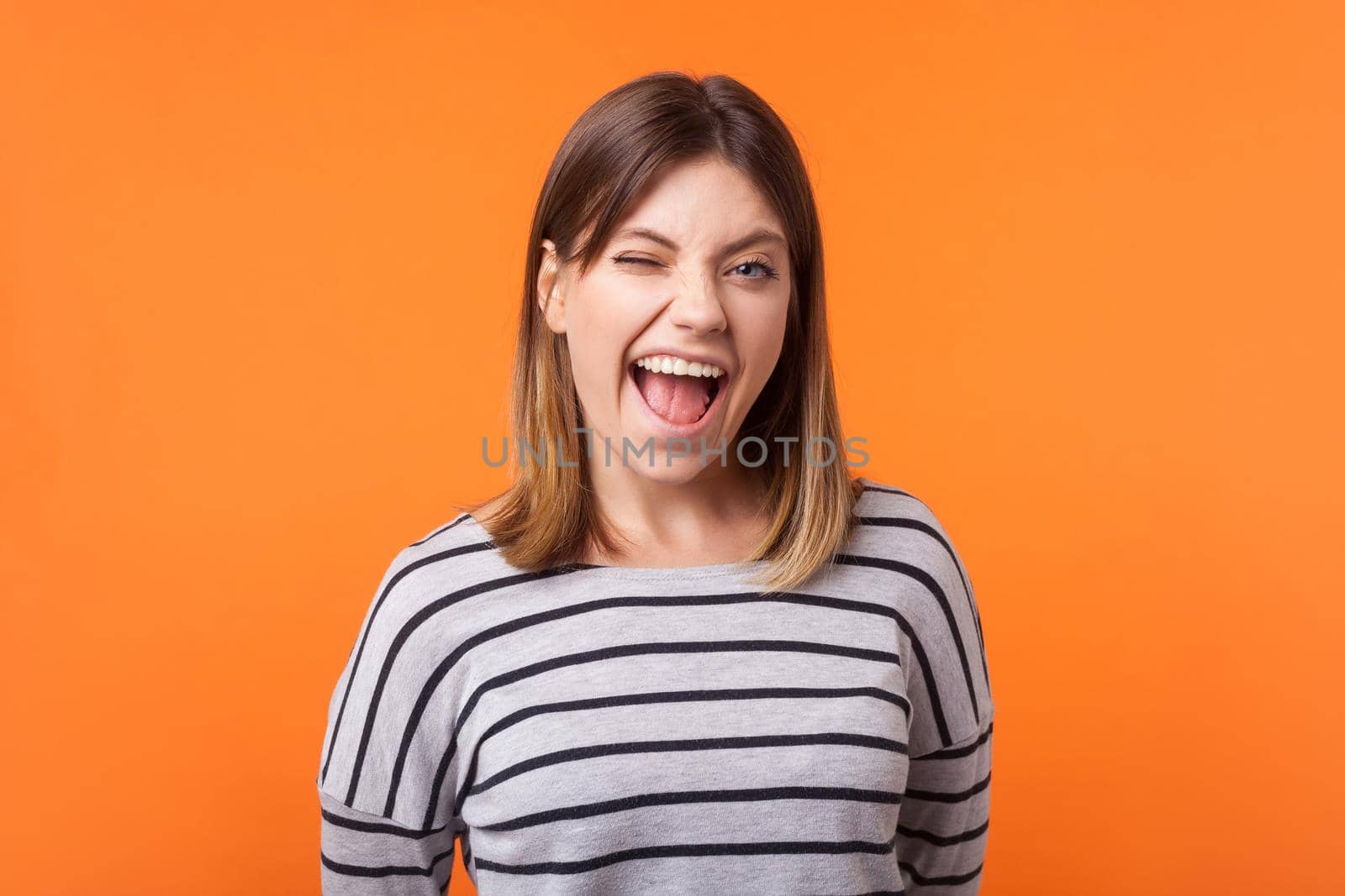 Portrait of cheerful amazing young woman with brown hair in long sleeve striped shirt standing with open mouth, winking playfully at camera, flirting. indoor studio shot isolated on orange background