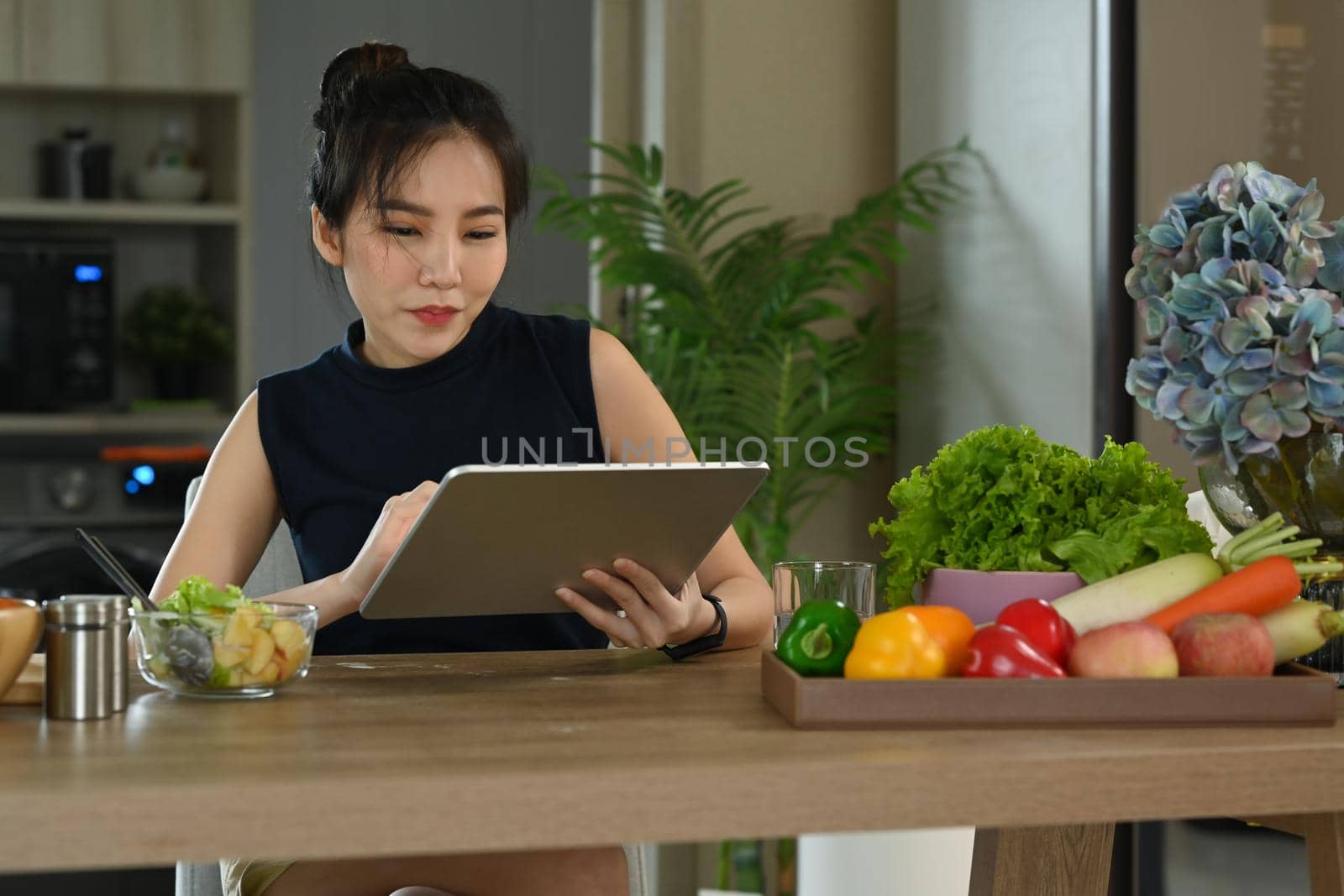 Young woman sitting in home kitchen and reading online recipe on digital tablet. by prathanchorruangsak