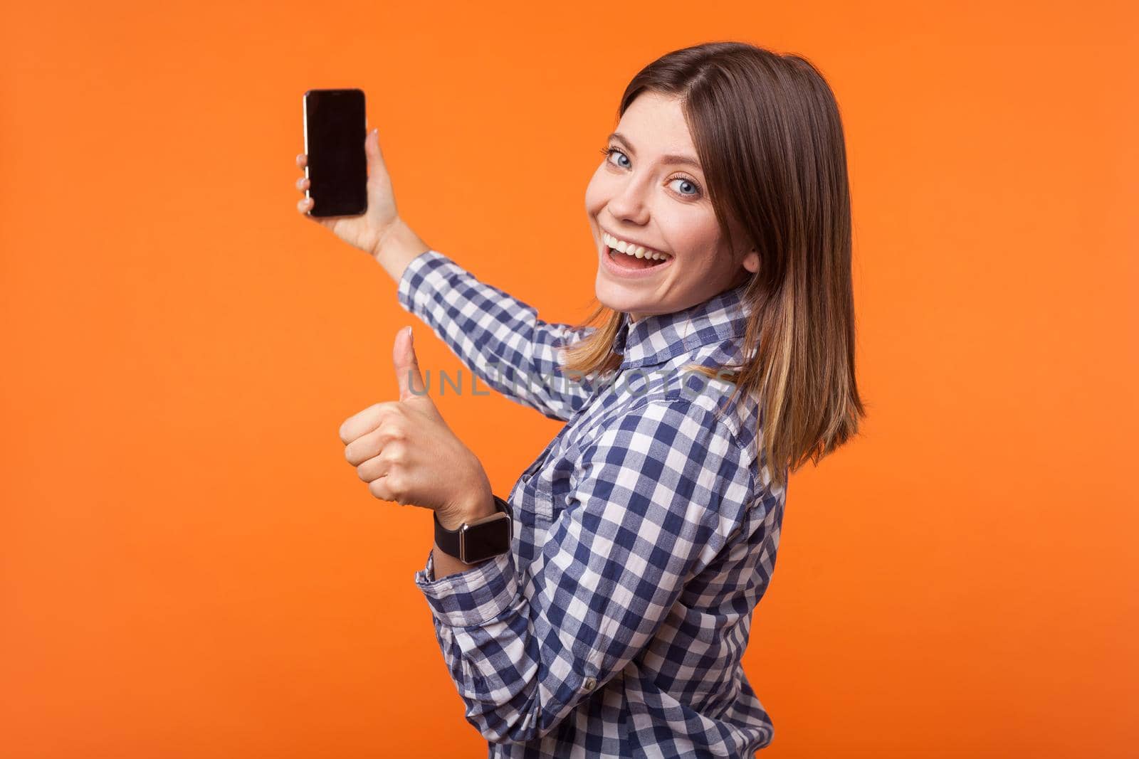 Half-turned portrait of brunette woman with charming smile in checkered shirt holding phone and showing thumbs up, satisfied with mobile services. indoor studio shot isolated on orange background