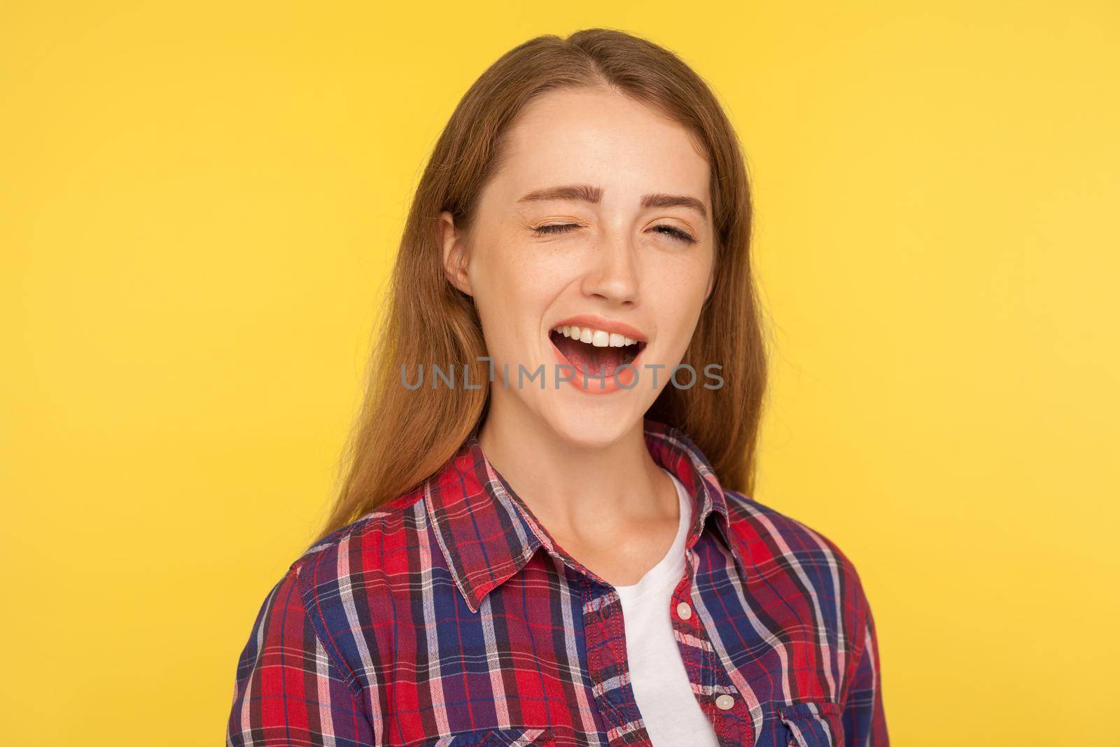 Portrait of cheerful optimistic ginger girl in checkered shirt winking at camera with pleasure, having playful happy expression, blinking eye and flirting. studio shot isolated on yellow background