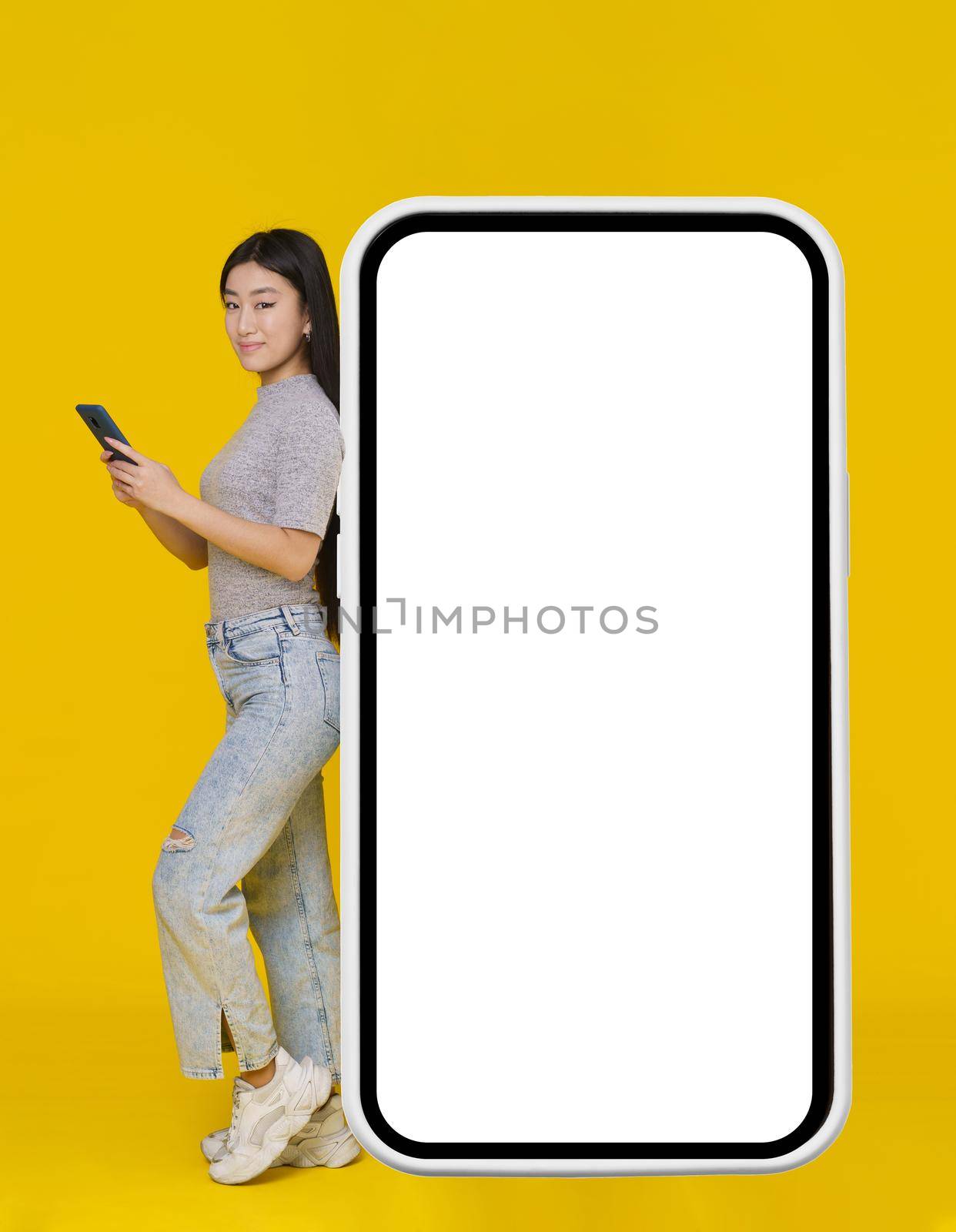 Smiling asian girl with phone in hands leaned back on giant, huge smartphone, digital tablet with white, black screen standing wearing casual isolated on yellow background. Free space mock up.