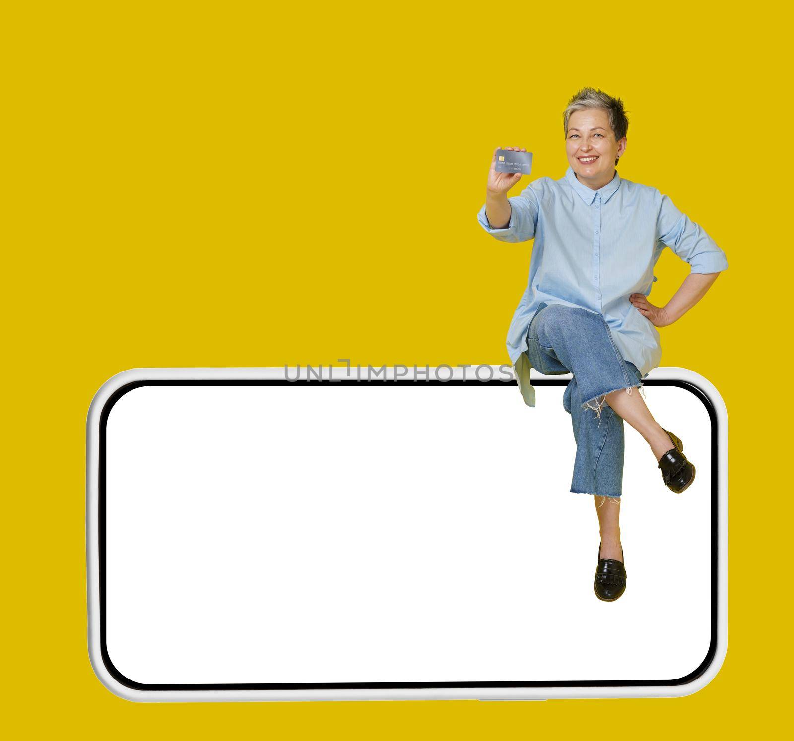 Happy grey haired mature woman sitting on a giant, huge smartphone, in casual blue shirt and jeans with hand raised up in a win, isolated on yellow background by LipikStockMedia