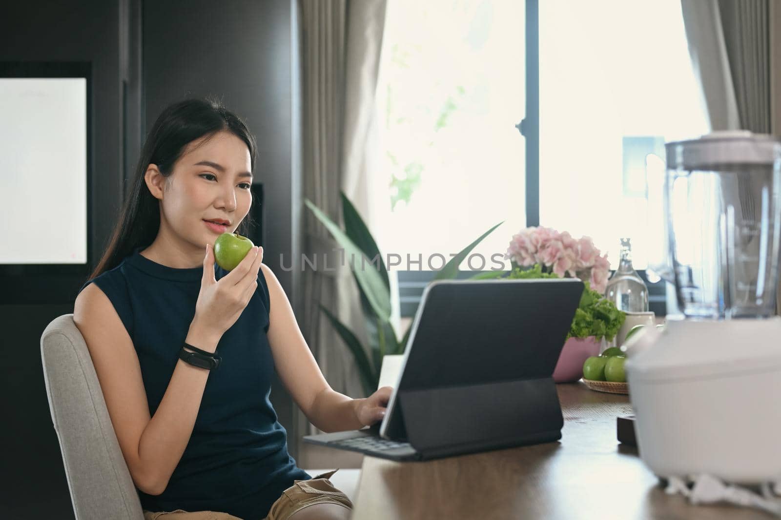 Healthy young woman eating apple and using computer tablet in kitchen.