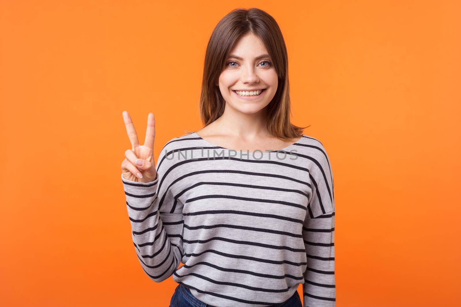 Portrait of happy charming woman with brown hair in long sleeve striped shirt smiling looking at camera and showing peace or victory sign with fingers. indoor studio shot isolated on orange background