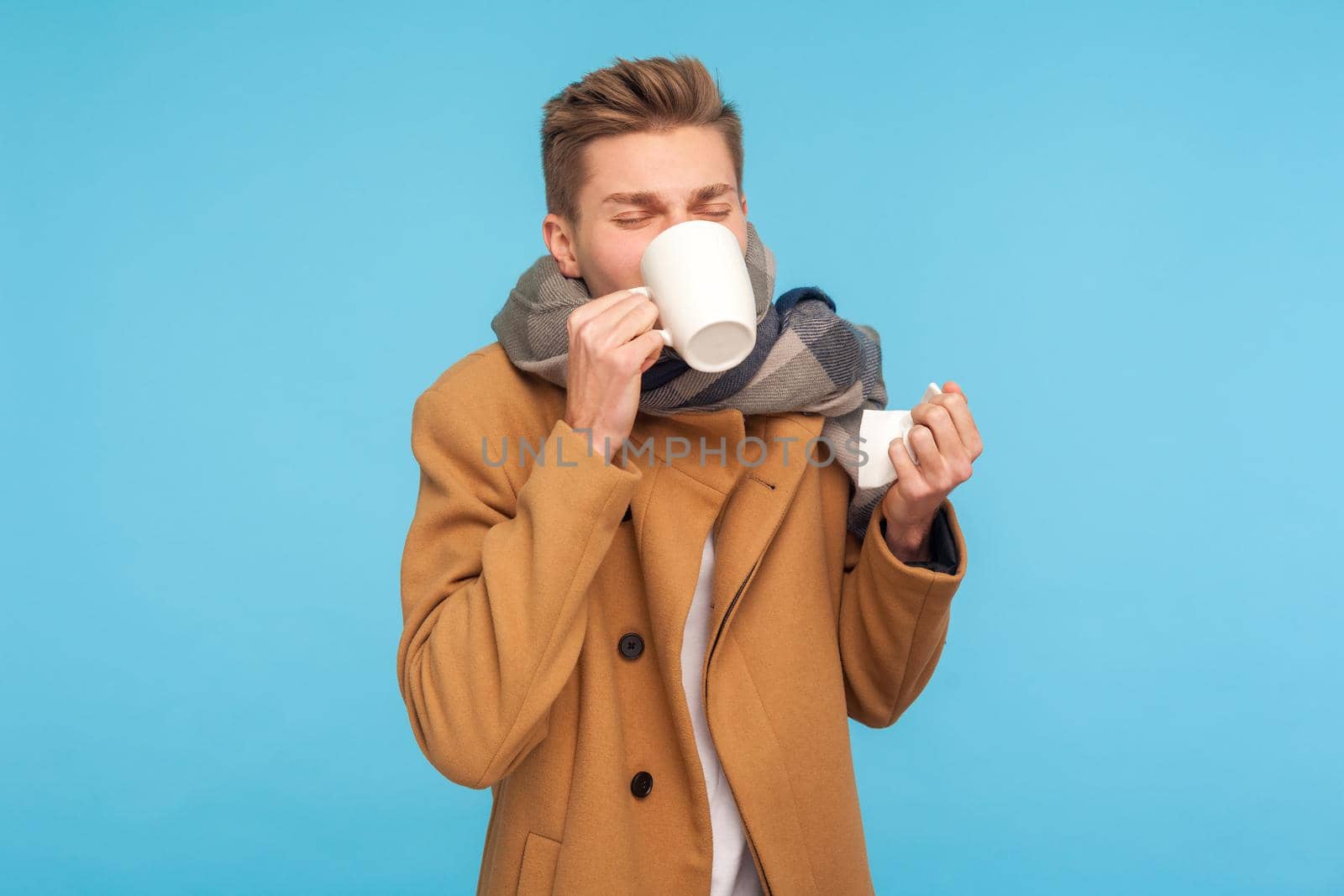 Flu treatment and self-care. Sick man in autumn coat and scarf drinking medicine with hot tea, holding tissue for runny nose, suffering fever, headache. indoor studio shot isolated on blue background