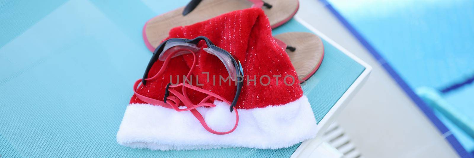 Hat of Santa Claus, swimming slippers on sunbed. Holidays near sea pool for Christmas holidays and relaxation, leisure, tourism concept.