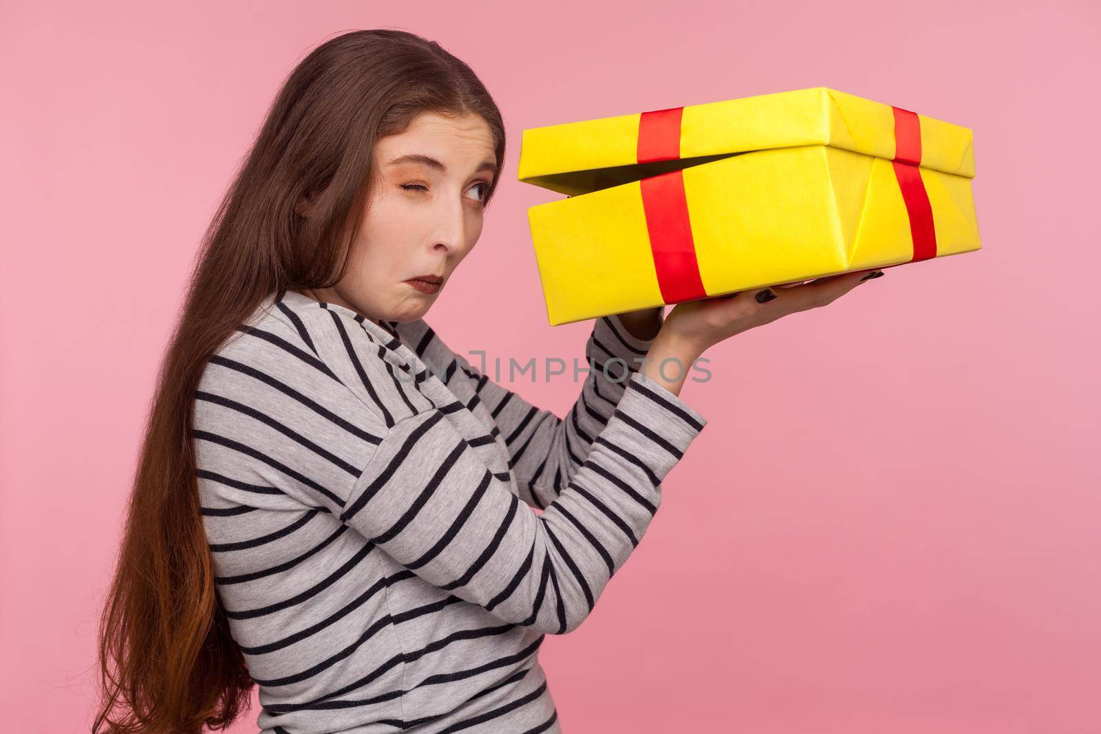 Portrait of funny curious nosy woman in striped sweatshirt peeking into gift box, impatient to unpack birthday present, looking inside with inquisitive expression. indoor studio shot, pink background