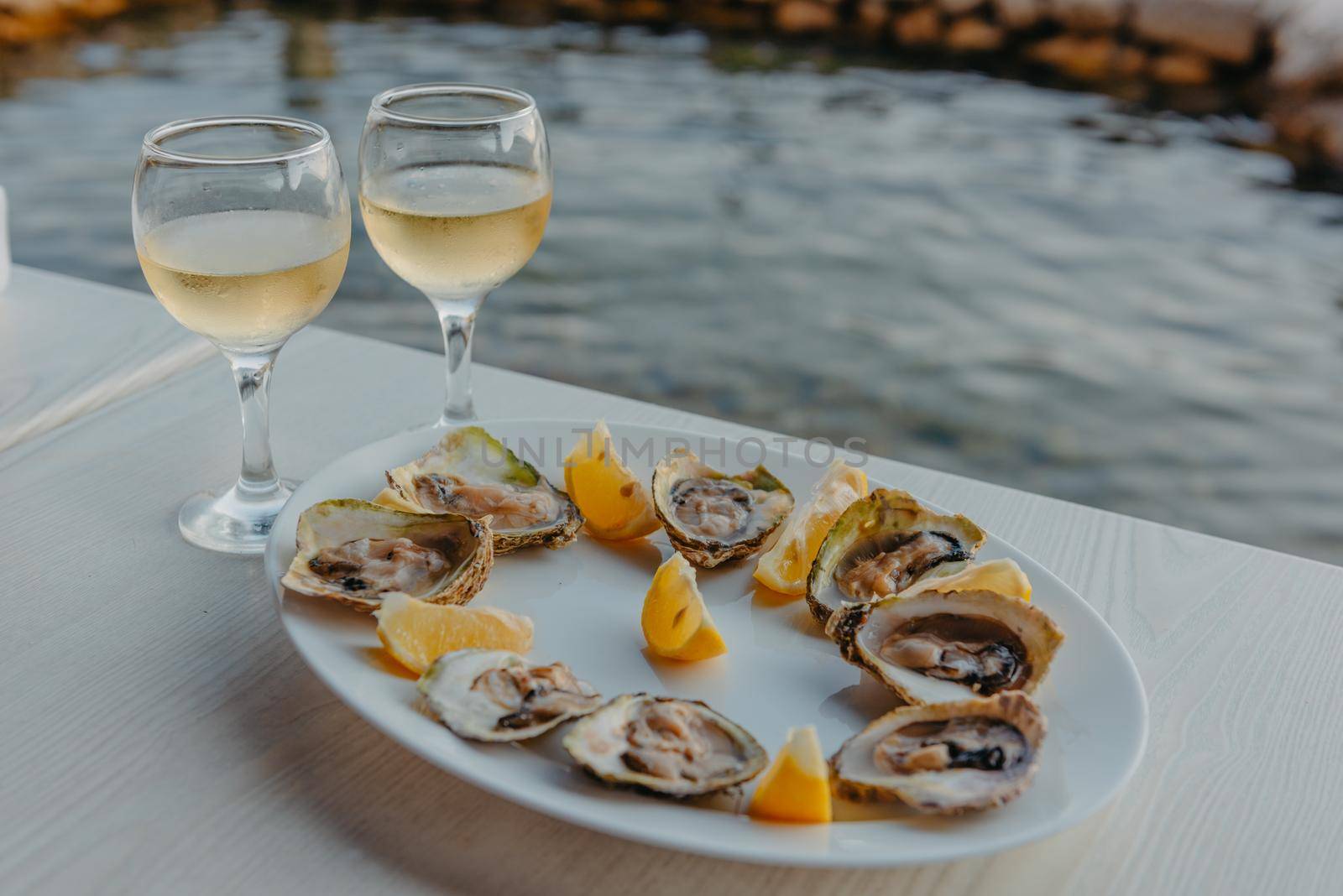 Freshly caught oysters on a plate and vine glasses. Restaurant on the shores of the Bay of Kotor near the oyster farm, Montenegro. Seafood. Beautuful seascape with oyster farm and mountains, Adriatic landscape, Montenegro. Oysters farm in sunset, Boka -Kotor Bay, Montenegro by Andrii_Ko