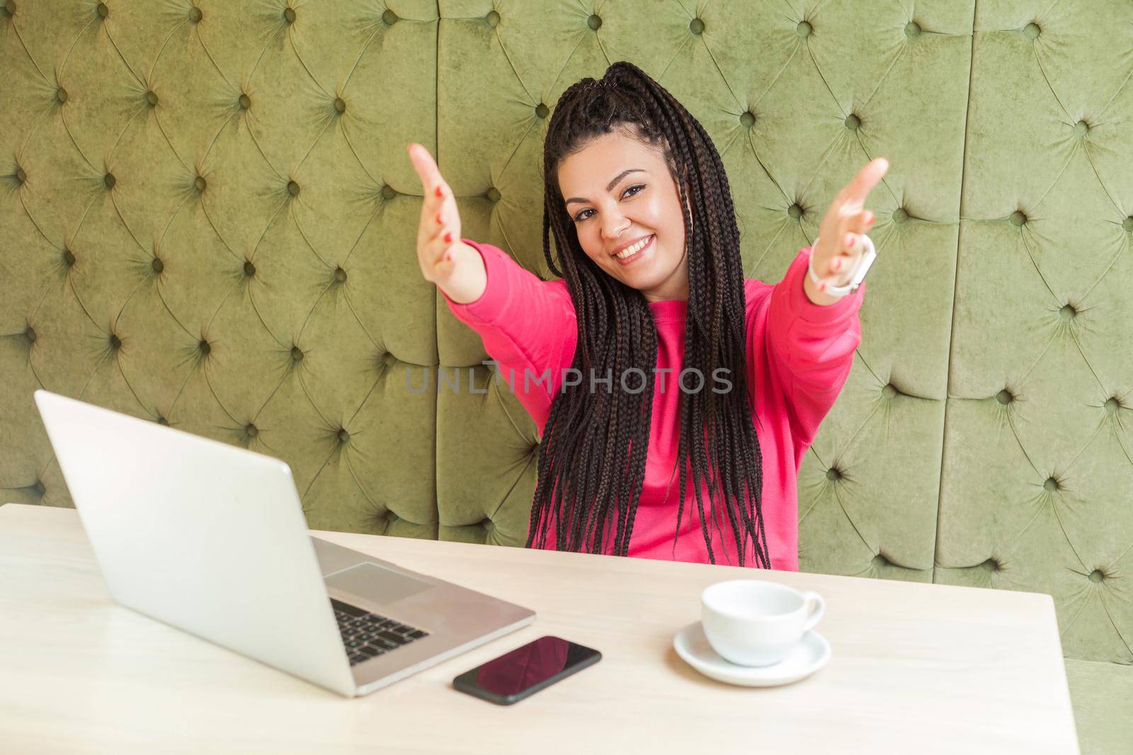 Portrait of lovely romantic young girl freelancer with black dreadlocks hairstyle are sitting alone in cafe and are ready hugging you with raised arms, toothy smile. Indoor, looking at camera
