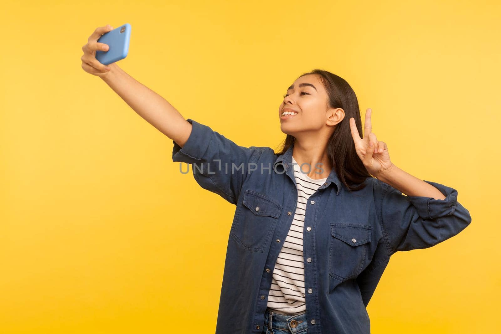 Portrait of successful stylish girl in denim shirt taking selfie or talking on video call and showing victory, peace gesture, pleasant online communication via mobile phone app. indoor studio shot