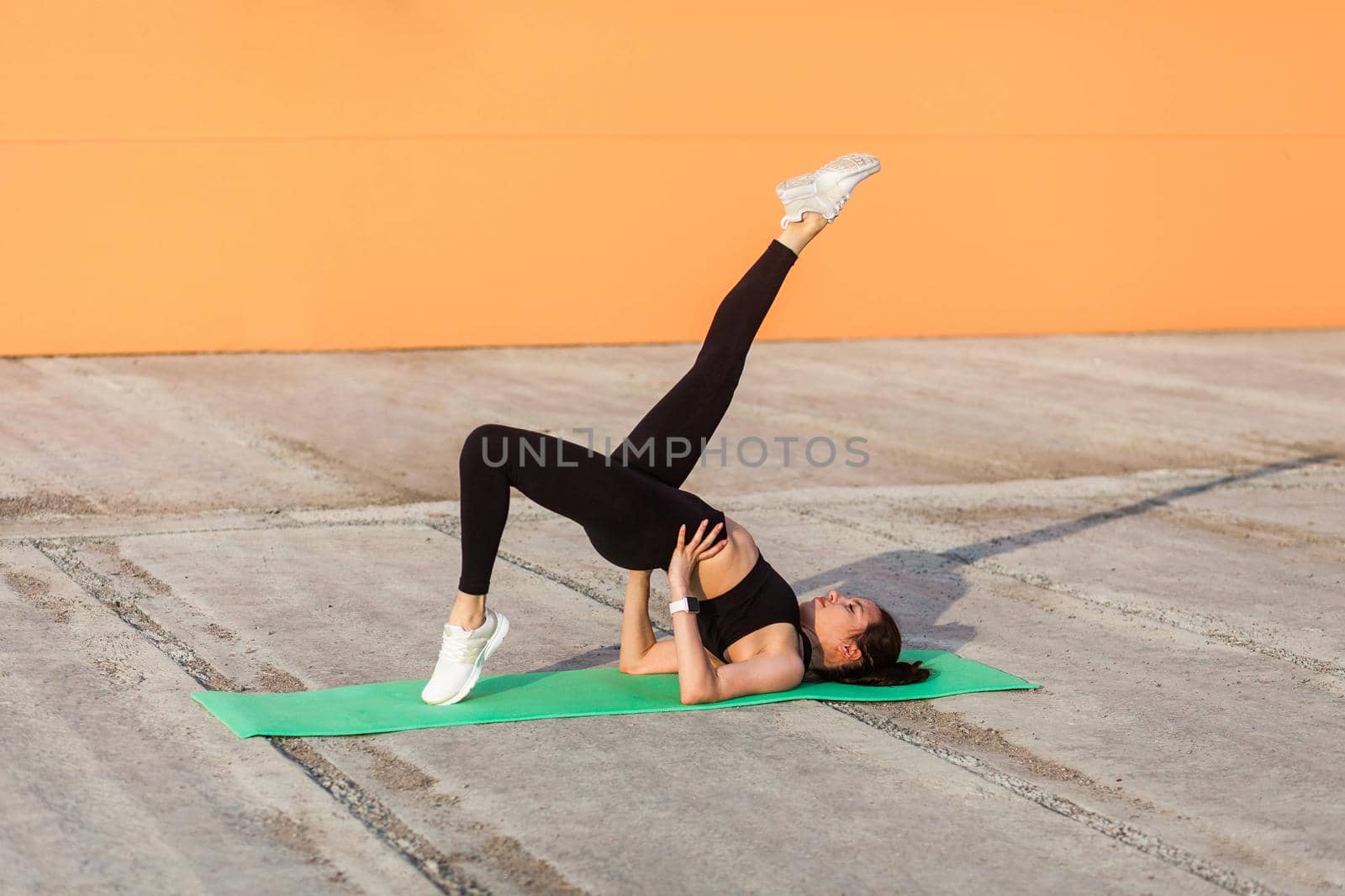 Slim athletic girl in tight sportswear, black pants and top, practicing yoga, doing one legged bridge pose with leg raise, training muscles and flexibility. Health care, sport activity outdoor