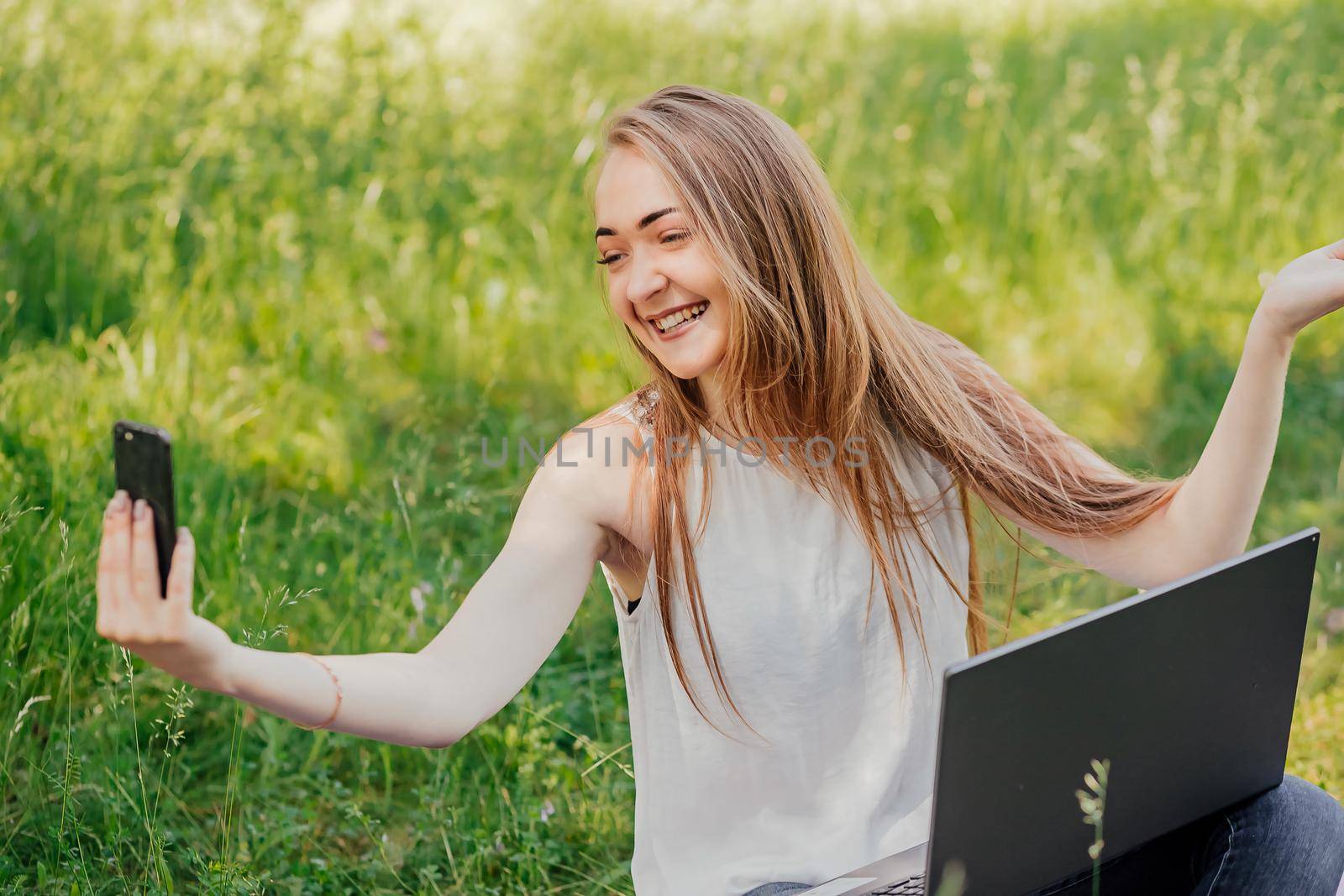 girl sits outdoors and works at a laptop. makes a video call on the phone. freelance. selfeducation. the concept of remote learning and outdoor work. by Anyatachka