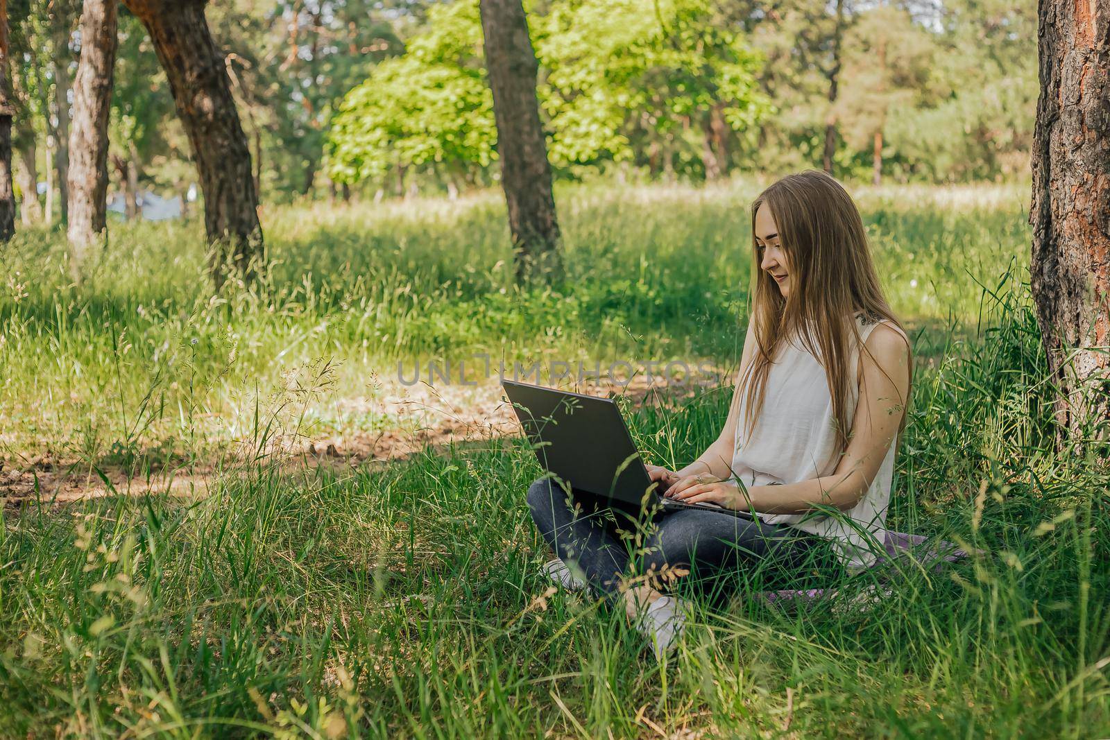 On the banner, a young girl works with a laptop in the fresh air in the park, sitting on the lawn. The concept of remote work. Work as a freelancer. The girl takes courses on a laptop and smiles