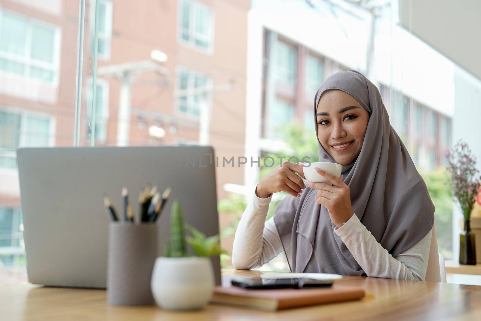 Attractive muslin woman working in office with laptop computer, hand holding cup of coffee