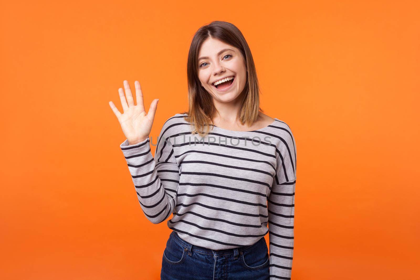 Hello. Portrait of adorable friendly woman with brown hair in long sleeve shirt standing waving hand, looking at camera with engaging toothy smile. indoor studio shot isolated on orange background