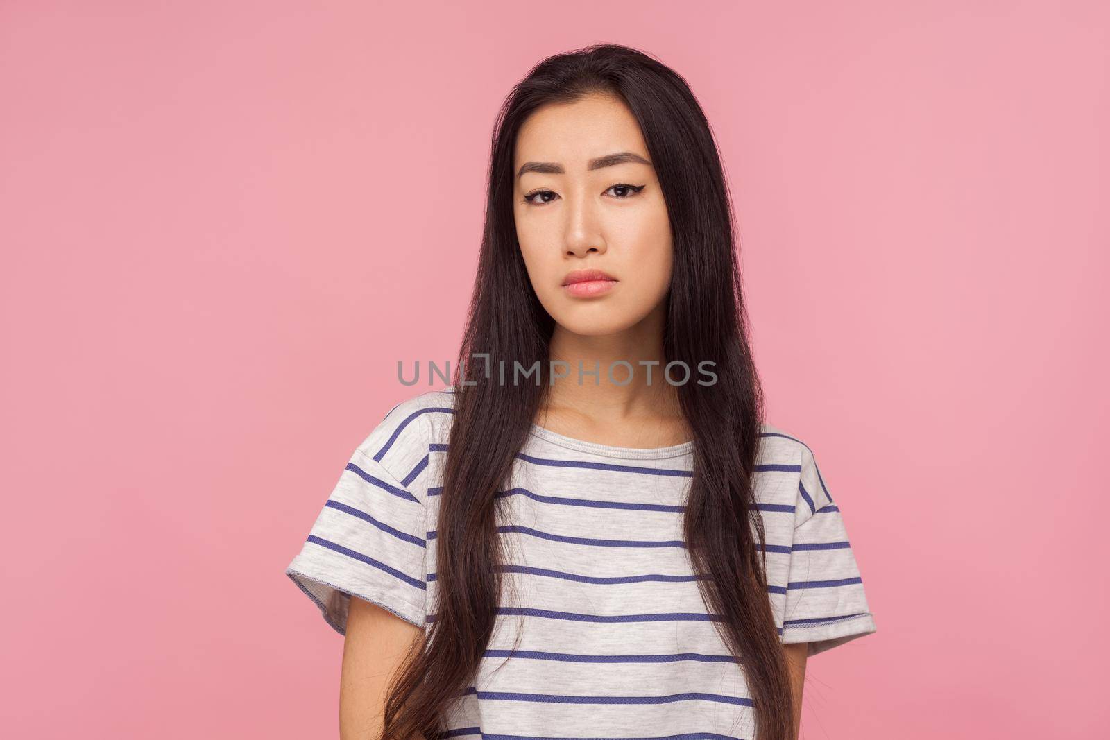 Portrait of sad unsmiling asian girl with long hair in striped t-shirt feeling frustrated resentful, looking dissatisfied with defeat, life troubles. indoor studio shot isolated on pink background