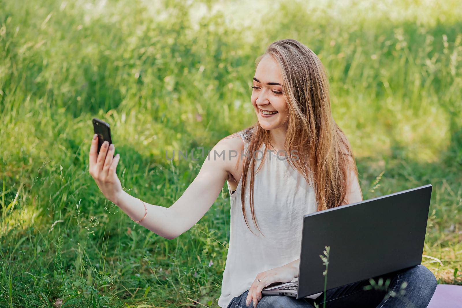 girl sits outdoors and works at a laptop. makes a video call on the phone. freelance. selfeducation. the concept of remote learning and outdoor work. by Anyatachka