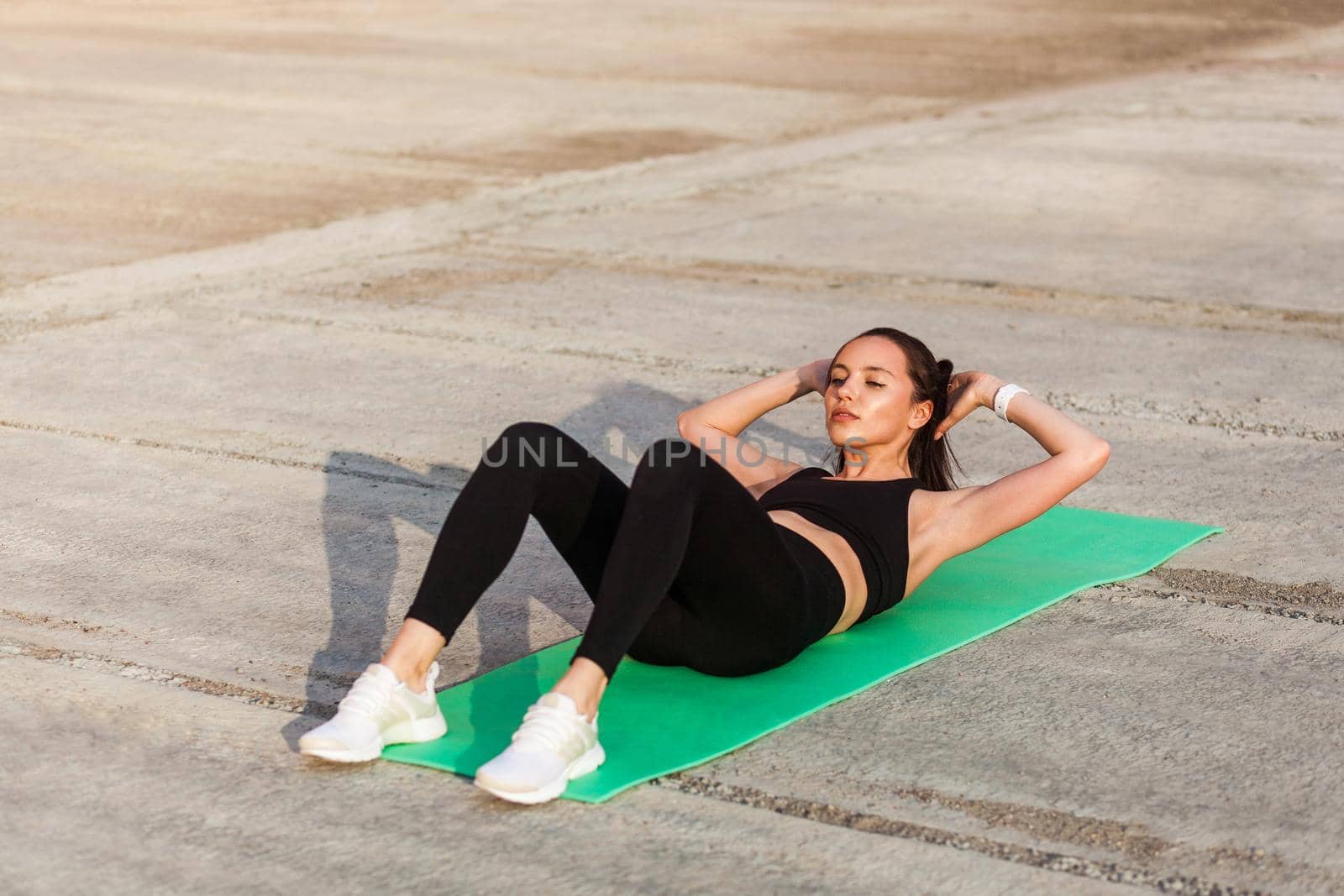 Athletic fit young woman in tight sportswear, black pants and top, lying on mat doing sit ups, crunch exercise, training abdominal muscles. Health care and weight loss concept, sport activity outdoor