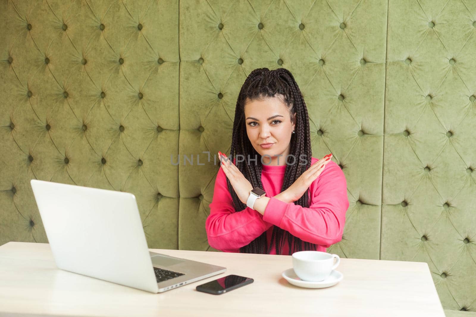 Warning aggressive young girl with black dreadlocks in pink blouse are sitting in cafe and working on laptop, showing crossing raised arms like stop or end gesture, looking at camera. Indoor