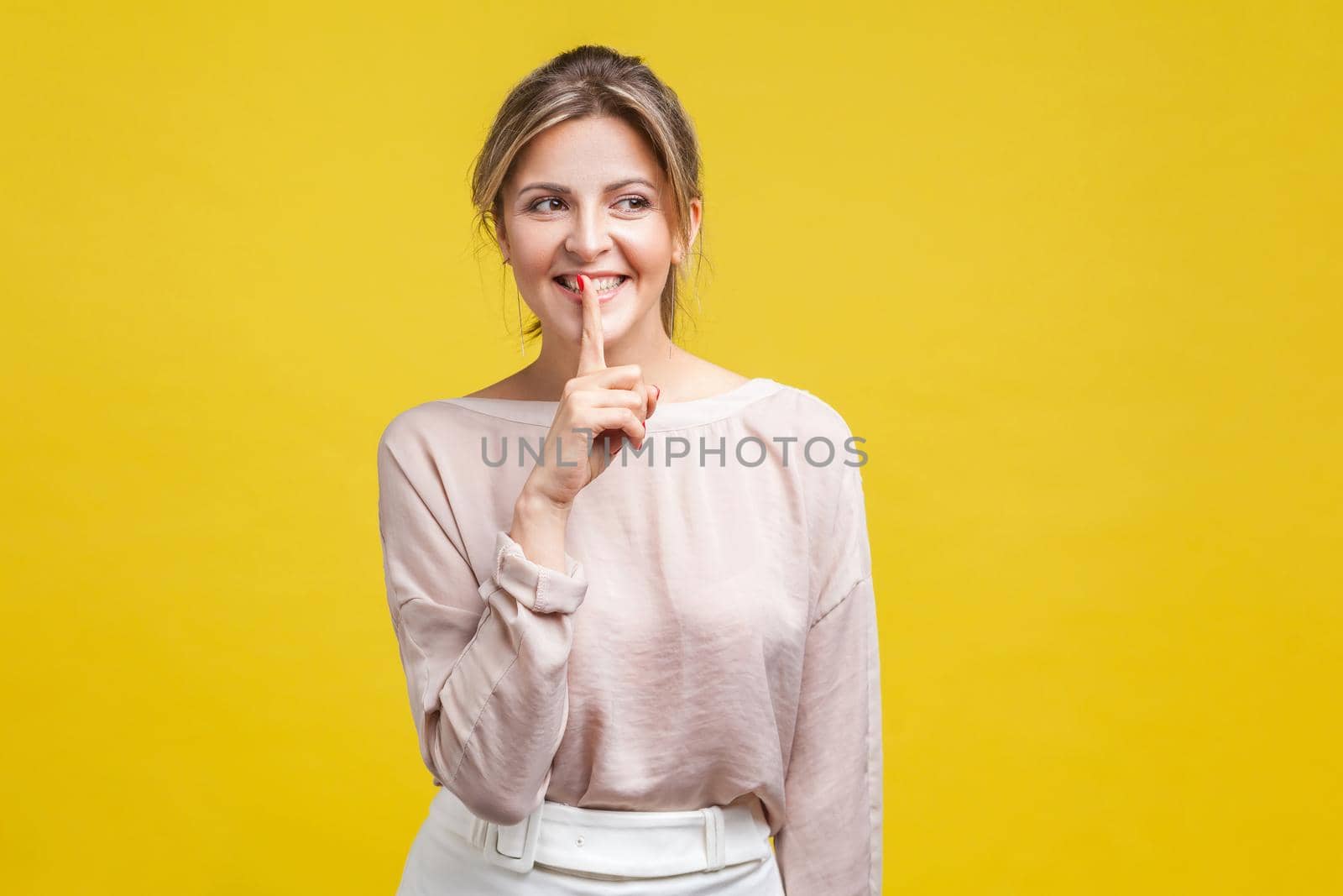 Portrait of cheerful beautiful girl with blonde hair in casual beige blouse standing, looking away with toothy smile and showing silence gesture, indoor studio shot, isolated on yellow background