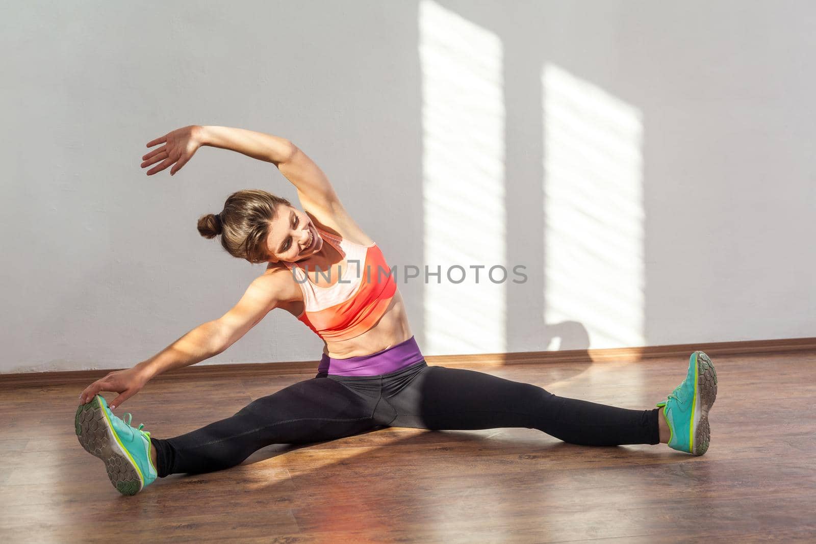 Beginner doing workouts. cheerful woman with bun hairstyle and in sportswear trying to do side bends while sitting on floor, practicing at home. indoor studio shot illuminated by sunlight from window