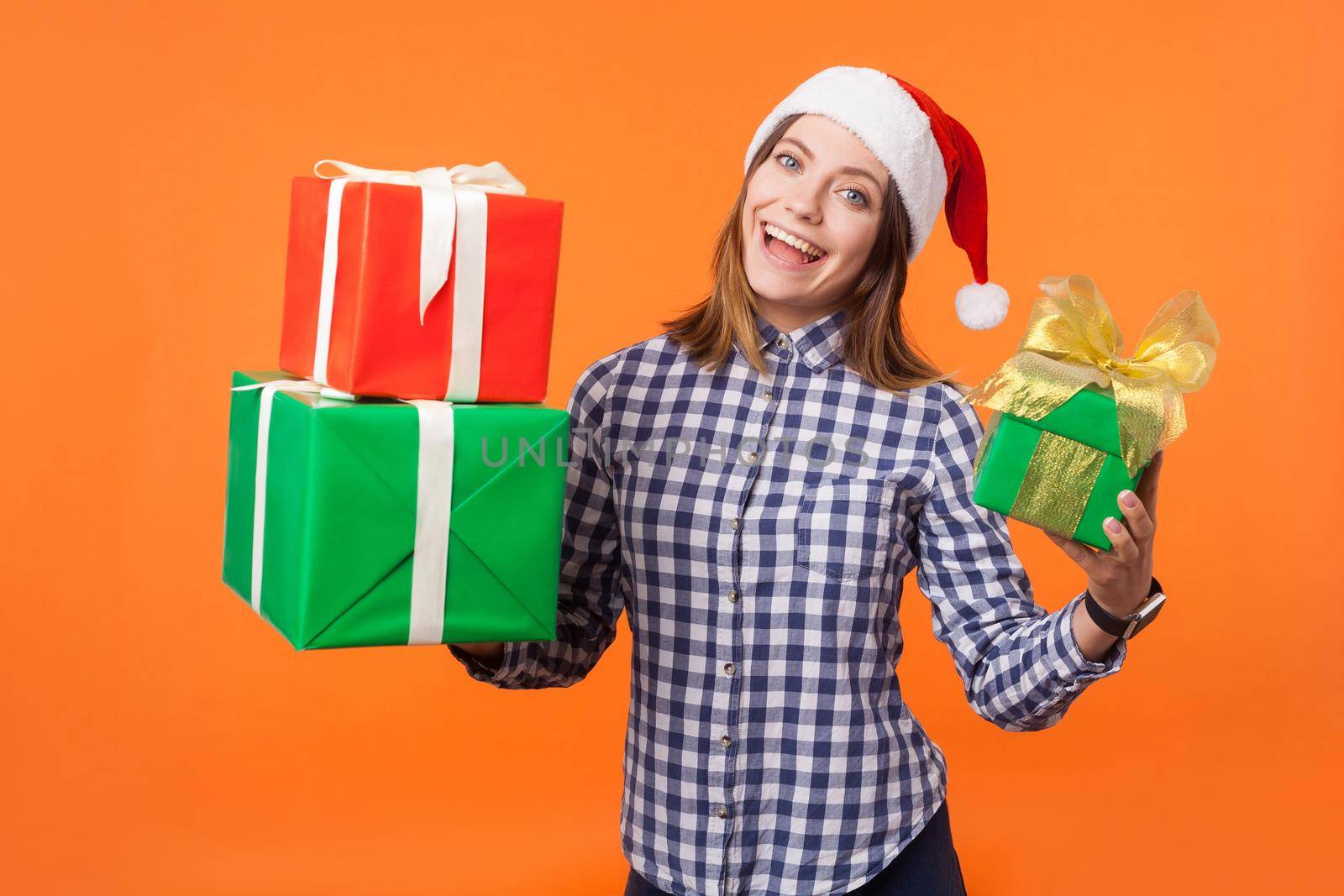 Portrait of joyous beautiful brunette woman in santa hat and checkered shirt standing, holding gift boxes and looking at camera with excitement. indoor studio shot isolated on orange background